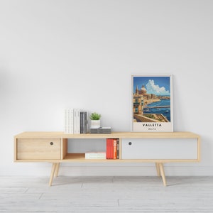 May include: A light wood entertainment center with two drawers, open shelves, and a white door. The center has a poster of Valletta, Malta, on the top shelf, along with books and a potted plant.