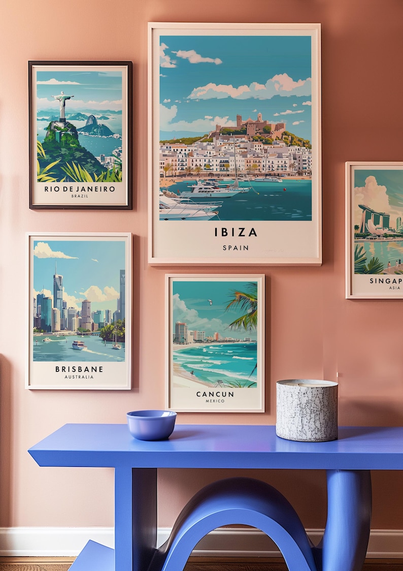 May include: Five framed travel posters with cityscapes and text below each image. The posters are hanging on a light pink wall. The posters are for Rio de Janeiro, Brazil; Ibiza, Spain; Brisbane, Australia; Cancun, Mexico; and Singapore, Asia. A blue table with a curved base is in the foreground.