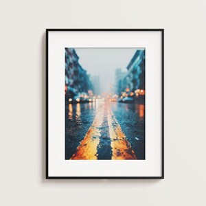 以下が含まれることがあります： 雨上がりの街並みを捉えた額装アートプリント。 アスファルトの道路には2本の黄色の線が中央にあり、街灯の温かい光を反射しています。通りの両側には建物が並び、背景はぼやけており、奥行きを演出しています。