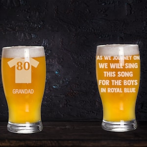 May include: Two clear glass pint glasses with a white design. The glass on the left has a yellow jersey with the number 80 and the word "GRANDAD". The glass on the right has the text "AS WE JOURNEY ON WE WILL SING THIS SONG FOR THE BOYS IN ROYAL BLUE".