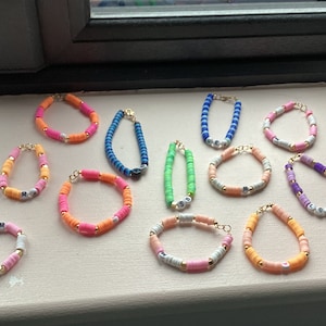 Könnte beinhalten: Eine Sammlung bunter Perlenarmbänder mit Buchstabenanhängern. Die Armbänder sind aus verschiedenen Farben von Perlen gefertigt, darunter Pink, Orange, Grün, Blau und Lila. Jedes Armband hat einen goldenen Verschluss und einen Buchstabenanhänger.