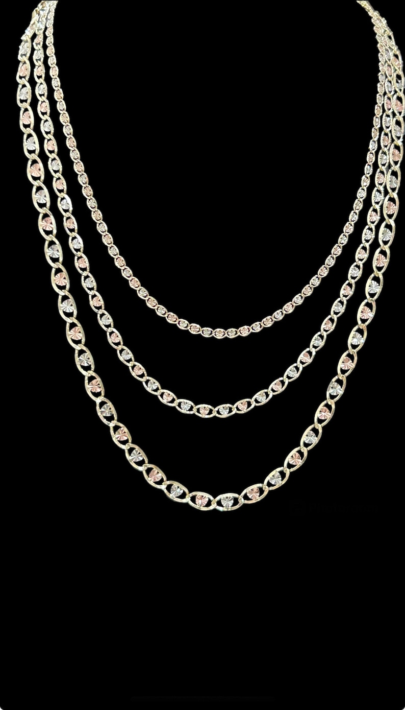 May include: Three gold chains with heart-shaped links. The chains are layered, with the largest chain on the bottom and the smallest chain on top. The chains are made of yellow, white, and rose gold.