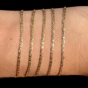 Pu&ograve; includere: Cinque collane a catena color oro con un design a maglie piatte e rettangolari. Le catene sono esposte su uno sfondo color carne, mostrando la loro delicata struttura e la calda tonalit&agrave;. Le catene sono probabilmente gioielli.