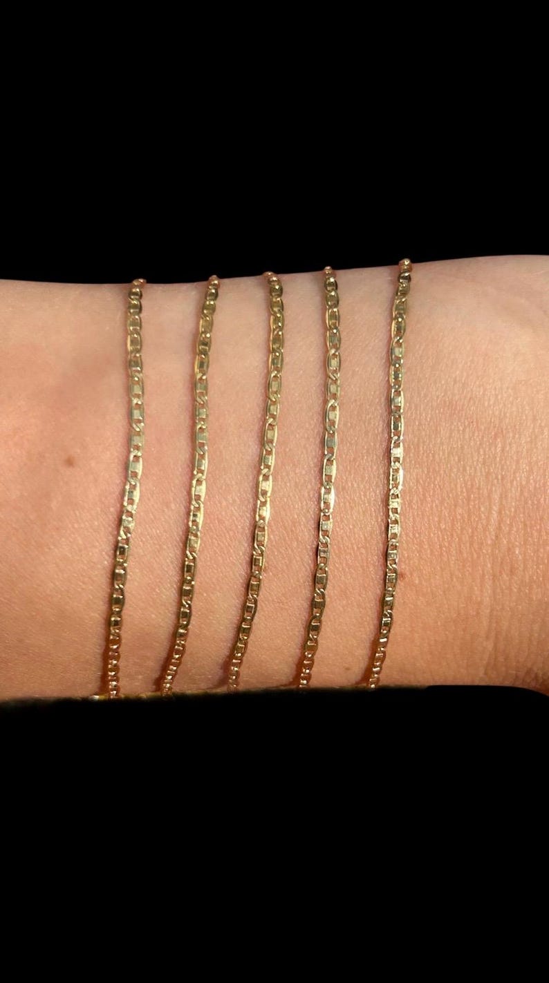 Pu&ograve; includere: Cinque collane a catena color oro con un design a maglie piatte e rettangolari. Le catene sono esposte su uno sfondo color carne, mostrando la loro delicata struttura e la calda tonalit&agrave;. Le catene sono probabilmente gioielli.