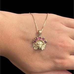 May include: A gold-colored pendant necklace featuring a Native American chief design. The pendant has a face with a headdress adorned with pink gemstones. The necklace is on a delicate gold chain, set against a dark background.