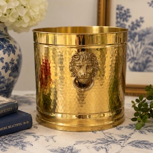 May include: A cylindrical brass bucket featuring a hammered texture and a prominent lion head handle on the front. The polished metal surface reflects light, highlighting the detailed relief of the lion's face and the attached circular ring.
