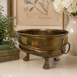 May include: An oval brass basin with a hammered texture, featuring four claw-shaped feet and two side handles adorned with lion head motifs. The metal has a dark, aged patina with visible surface variations and a wide, open top rim.