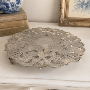 May include: A round silver-toned metal cake stand featuring an intricate openwork border with embossed scroll and floral patterns. The circular tray is supported by small, ornate feet, showcasing a metallic finish with detailed decorative cutouts.