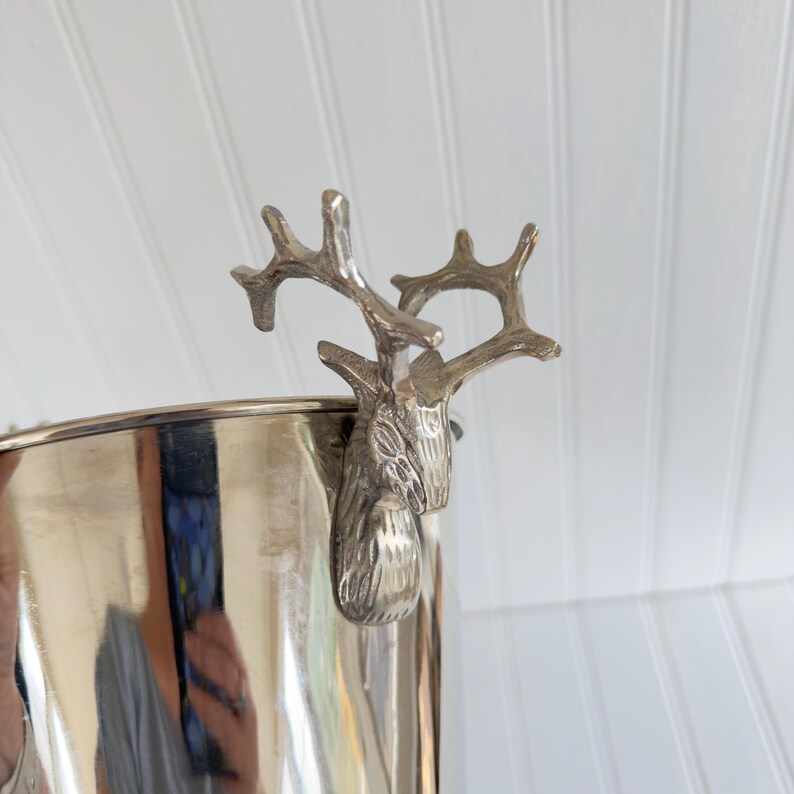 May include: A silver-colored metal ice bucket with a deer head and antler design on the side. The bucket has a reflective surface and a cylindrical shape. The deer head is detailed and adds a decorative touch to the barware.