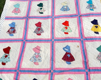 Puede incluir: Una colcha con una cuadrícula de cuadrados blancos separados por tiras de tela azul y rosa. Cada cuadrado contiene una figura aplicada que lleva un sombrero de ala ancha y un vestido hecho de varias telas estampadas en colores rojo, azul, verde y amarillo.