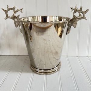 May include: A silver-colored metal ice bucket with a flared shape and decorative deer antler handles. The bucket has a ridged design on the exterior and a circular base. The handles are detailed with the shape of deer antlers.