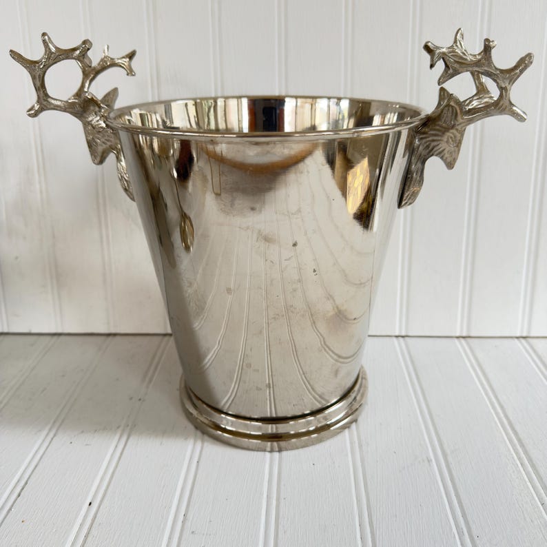 May include: A silver-colored metal ice bucket with a flared shape and decorative deer antler handles. The bucket has a ridged design on the exterior and a circular base. The handles are detailed with the shape of deer antlers.