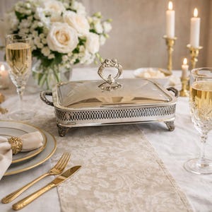 Op de afbeelding: Een zilveren rechthoekige serveerschaal met een glazen deksel, sierlijke handgreep en decoratieve opengewerkte rand. De schaal staat op een tafel gedekt voor een formele gelegenheid, met champagneglazen, kaarsen en een bloemstuk.