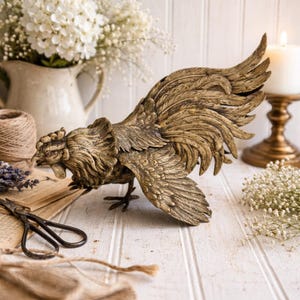 May include: A detailed bronze-colored rooster sculpture with outstretched wings and open beak. The sculpture is on a white wooden surface, with a blurred background of flowers, a candle, and vintage items.