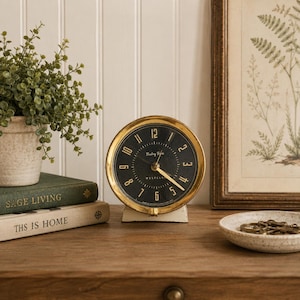 May include: A round Westclox Baby Ben alarm clock with a metallic gold-tone bezel and a black face featuring gold-colored numerals and hands. The clock is mounted on a small, off-white base and displays the brand name Westclox on the dial.