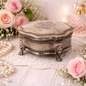 May include: A vintage silver jewelry box with ornate detailing, resting on a white lace doily. The box features a decorative lid and lion's paw feet. Pink roses and pearls surround the box, creating a romantic and elegant display.