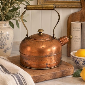 May include: A round copper tea kettle featuring a weathered patina, a brass handle, and a matching brass lid knob. The body of the kettle displays a subtle engraved floral band around the centre and a short spout with a small brass finial.