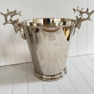 May include: Silver-colored metal wine bucket with a flared shape and a round base. The bucket features decorative deer antler handles on either side. The interior of the bucket is reflective, and the exterior has vertical etched lines.