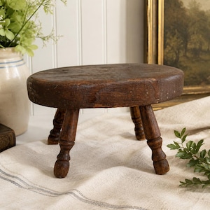 May include: A small, round, dark brown wooden stool featuring a smooth, weathered top surface and three turned legs. The stool has a visible wood grain texture and shows signs of age and wear, with a sturdy construction and a low profile design.