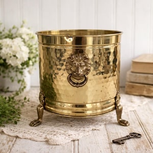Puede incluir: Una jardinera de latón martillado con un asa de anillo con cabeza de león y patas decorativas. La jardinera redonda tiene una superficie texturizada. Se ve una llave antigua. Esta jardinera decorativa es adecuada para uso en interiores o exteriores.