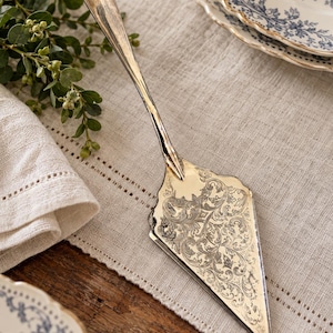 May include: A silver-toned metal cake server featuring an elongated handle and a triangular blade. The blade is decorated with intricate, engraved scrollwork patterns across its surface, while the edges of the blade are smooth and polished.