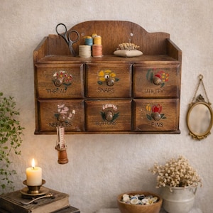 Puede incluir: Un organizador de costura de madera vintage con seis cajones etiquetados "Thread", "Snaps", "Utility", "Needles", "Buttons" y "Pins". Los cajones tienen detalles florales y pomos redondos. Tijeras, carretes de hilo y un alfiletero encima.