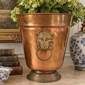 May include: A large, tapered copper planter with a weathered patina, featuring a prominent brass lion head ornament on the front. The lion head holds a circular brass ring in its mouth. The planter has a wide, flared rim and a circular base.