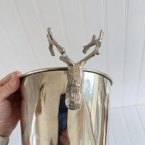May include: A silver-colored metal ice bucket with a decorative deer head handle. The bucket has a polished finish and a cylindrical shape. The deer head is detailed with antlers and a textured face.