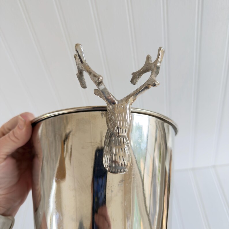 May include: A silver-colored metal ice bucket with a decorative deer head handle. The bucket has a polished finish and a cylindrical shape. The deer head is detailed with antlers and a textured face.