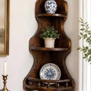Può includere: Una mensola angolare in legno marrone scuro a tre livelli. Il ripiano superiore contiene un vaso con coperchio blu e bianco. Il ripiano centrale presenta una pianta verde in un vaso chiaro. Il ripiano inferiore mostra un piatto con motivo floreale blu e bianco.