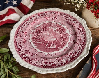 May include: A white and red decorative plate with a scalloped edge. The plate features a detailed red illustration of a landscape scene surrounded by a floral and leaf pattern. The plate is on a wooden surface.
