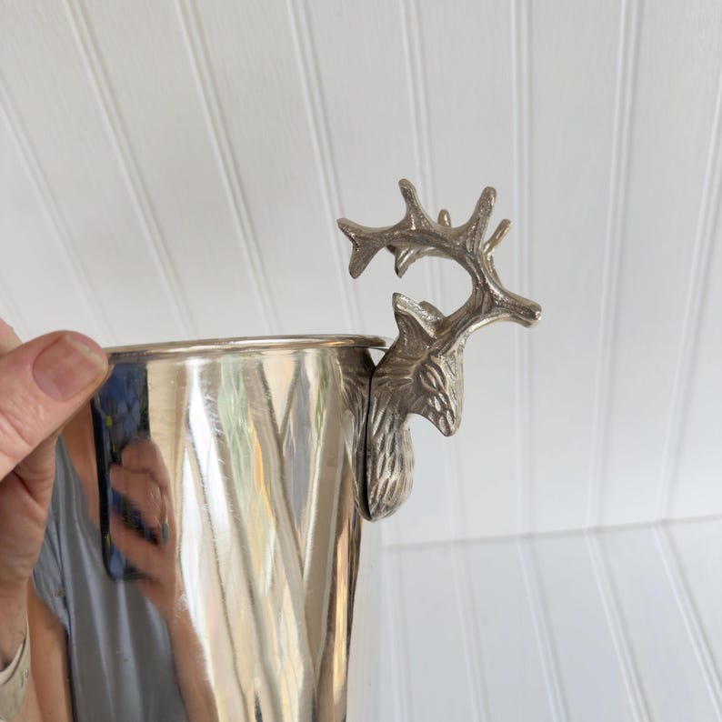 May include: A silver-colored metal cup with a decorative deer head handle. The cup has a smooth, reflective surface. The deer head is detailed with antlers and facial features. The cup is held by a person.