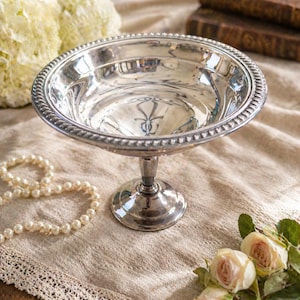 May include: A silver-colored pedestal bowl with a beaded rim. The bowl has a wide, shallow basin and a short, round stem. The bowl is sitting on a beige surface with flowers and a pearl necklace in the background.