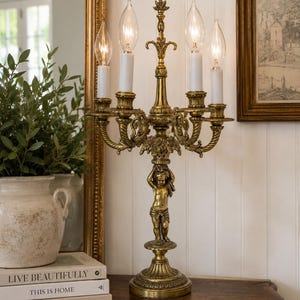 Puede incluir: Una lámpara candelabro de metal dorado con cuatro bombillas tipo vela y fundas blancas. El tallo central está sostenido por una figura esculpida de un querubín sobre una base circular, con detalles ornamentales de hojas y cuerdas.