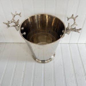 May include: A silver metal ice bucket with deer antler handles. The bucket has a tapered shape and a round base. The handles are detailed and add a rustic touch to the design. The interior of the bucket is reflective.