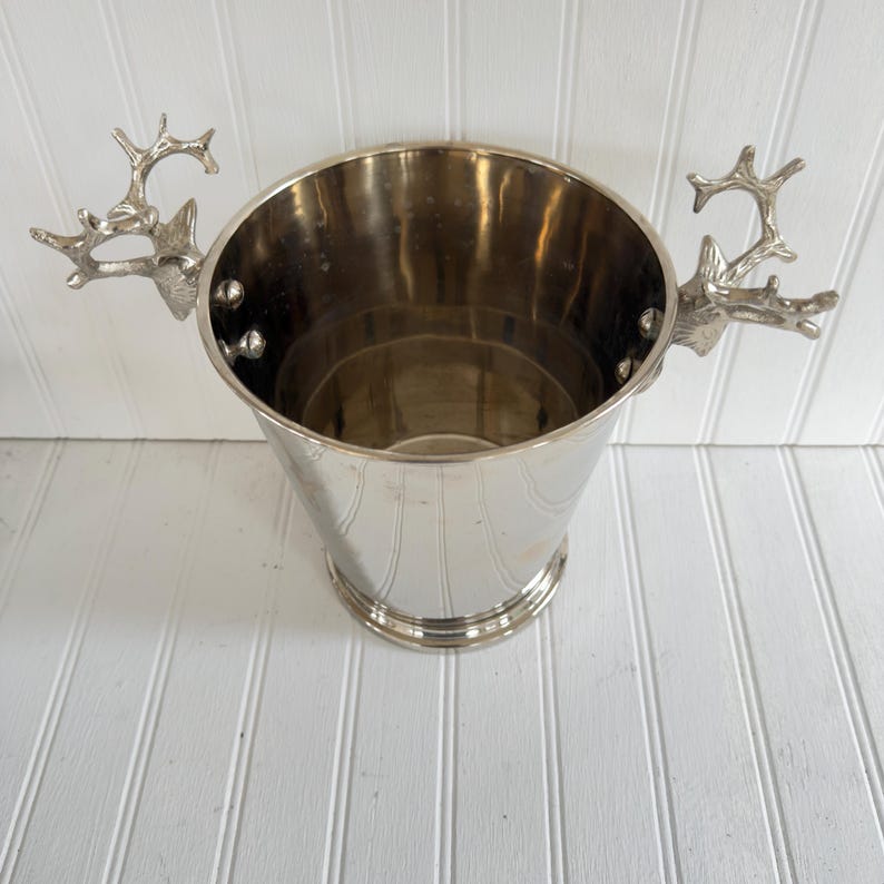 May include: A silver metal ice bucket with deer antler handles. The bucket has a tapered shape and a round base. The handles are detailed and add a rustic touch to the design. The interior of the bucket is reflective.