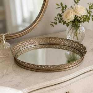 Puede incluir: Una bandeja decorativa redonda con base de espejo y borde metálico en tono dorado que presenta un intrincado patrón de celosía trenzada. La bandeja tiene un perfil bajo y una superficie reflectante pulida para colocar objetos.