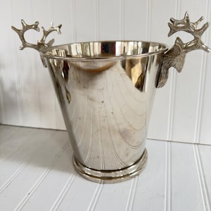 May include: Silver-colored metal ice bucket with deer antler handles. The bucket has a tapered shape and a wide, circular base. The antlers are detailed and add a rustic touch. The bucket is designed to hold ice and chill beverages.