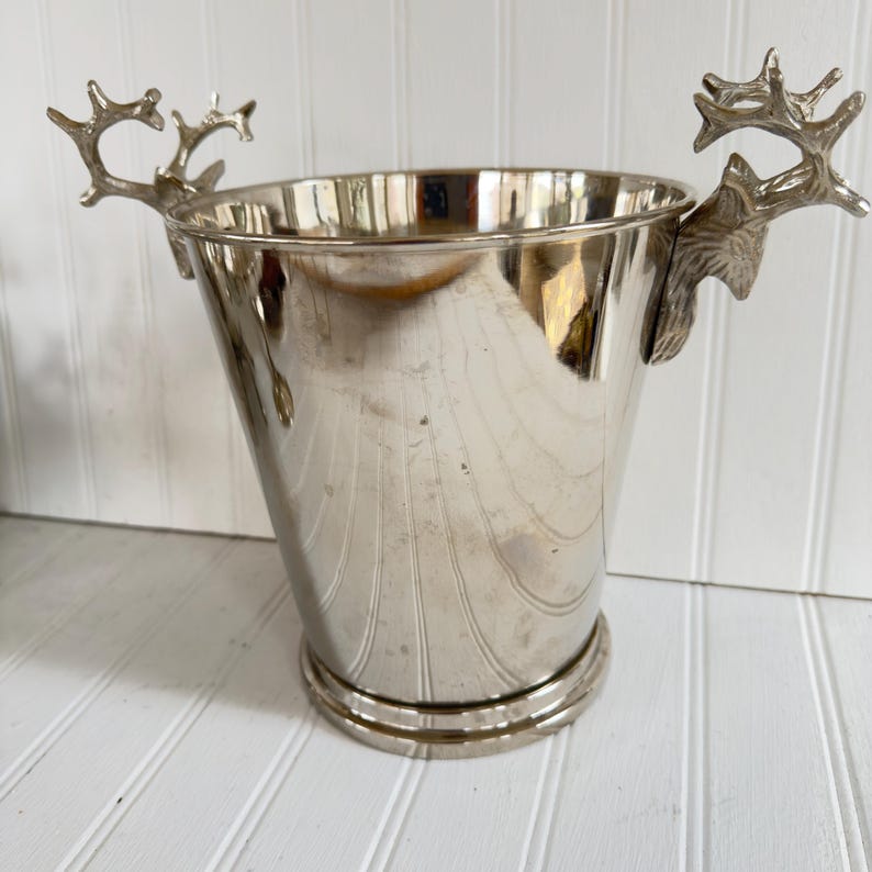 May include: Silver-colored metal ice bucket with deer antler handles. The bucket has a tapered shape and a wide, circular base. The antlers are detailed and add a rustic touch. The bucket is designed to hold ice and chill beverages.