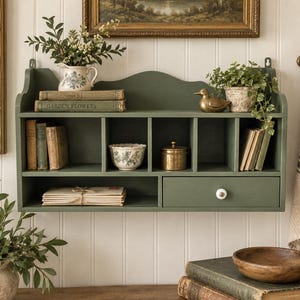 May include: A wall-mounted sage green wooden shelf unit featuring multiple open cubbies, a single drawer with a white knob, and a scalloped top edge. The shelf holds books, a small pitcher, a bowl, a brass container, and a decorative duck.