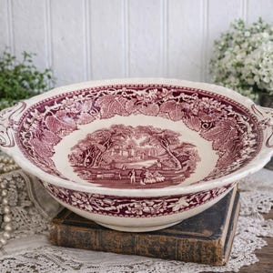 May include: A large, antique, cream-colored ceramic bowl with a burgundy landscape design. The bowl has a scalloped edge and handles. The interior features a detailed scene of people and buildings. The bowl is resting on a stack of old books.