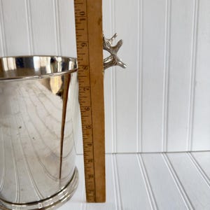 May include: A silver-colored metal ice bucket with a decorative handle. The bucket is approximately 8 inches tall, as measured by a wooden ruler. The bucket has a curved design on the side.