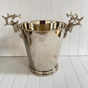 May include: A silver-colored metal ice bucket with a flared shape and decorative vertical lines. The bucket features handles shaped like deer antlers on either side. The bucket sits on a round base.