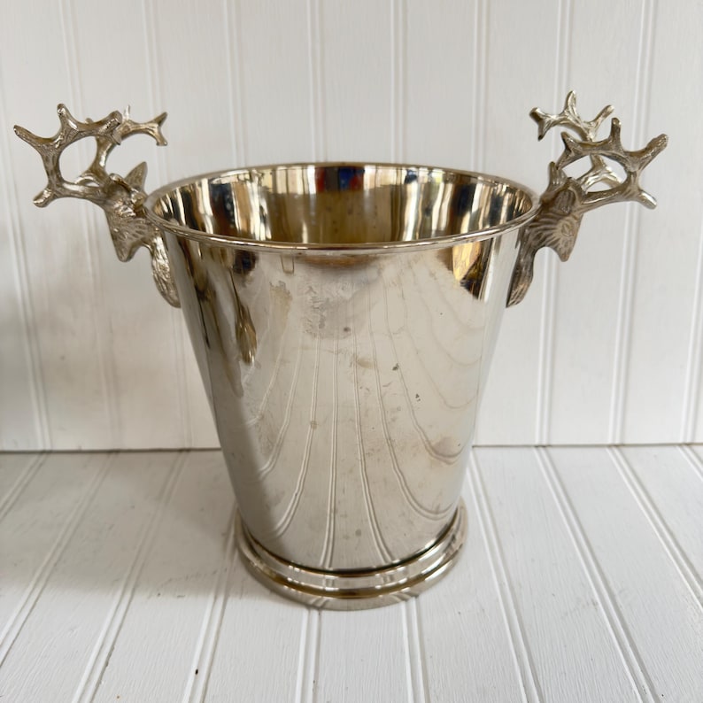 May include: A silver-colored metal ice bucket with a flared shape and decorative vertical lines. The bucket features handles shaped like deer antlers on either side. The bucket sits on a round base.