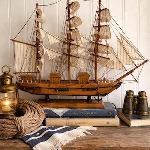 May include: A detailed wooden model ship with sails, displayed on a wooden table. The ship is brown with white sails and rigging. A lit brass lantern, rope, books, and binoculars complete the nautical theme.