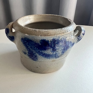 Könnte beinhalten: Ein weißer Keramiktopf mit blauer Glasur und zwei Henkeln. Der Topf hat eine raue, strukturierte Oberfläche.