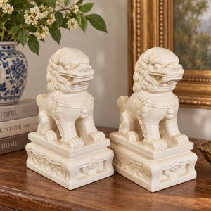 May include: A pair of off-white ceramic Foo Dog statues, each featuring intricate sculpted manes, snarling expressions, and detailed collars. The figures are mounted on rectangular pedestals with carved floral motifs on the front and sides.