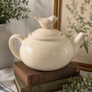 May include: A cream-colored ceramic teapot with a rounded body, a curved handle, and a spout. The lid features a sculpted bird finial with a distressed finish. The teapot has a smooth, glossy glaze and sits on a stack of two vintage hardcover books.