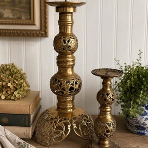 May include: Two brass candle holders with intricate openwork filigree patterns. Each holder features a central stem with spherical sections and a wide, ornate base with cut-out floral designs. The metal has a polished, aged gold-toned finish.