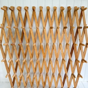 May include: A natural wood, concertina-style wall rack with multiple pegs for hanging items. The rack is made of light-coloured wood and has a diamond-shaped design when fully extended. It is mounted on a white, vertical-panelled wall.